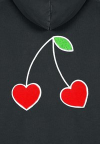 Due ciliegie a forma di cuore rosse con una foglia verde su un tessuto nero, posizionate sotto una cucitura del cappuccio.