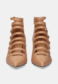 Zapatos de tacón alto de punta en cuero marrón claro con múltiples correas horizontales y botones redondos decorativos. Cierre con cremallera lateral.