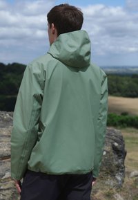 Hellgrüne Kapuzenjacke mit glatter Textur und elastischen Bündchen, für eine lockere Passform entworfen, von hinten vor natürlichem Hintergrund gezeigt.