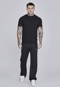 Schwarzes, tailliertes T-Shirt mit goldener Schrift auf den Ärmeln, kombiniert mit schwarzen Hosen mit einem goldenen Logo. Glatter Stoff und athletisches Design.