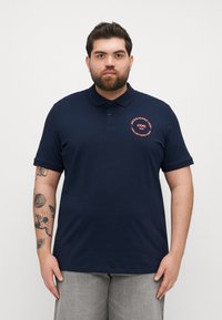 Polo din bumbac, de culoare bleumarin, cu guler clasic, mâneci scurte și un logo circular pe piept, de culoare coral contrastantă.