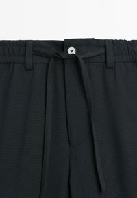 Sort bukseband med elastik, snøre bundet i en sløjfe, bæltestropper og en lille sort knap over lynlåsen.