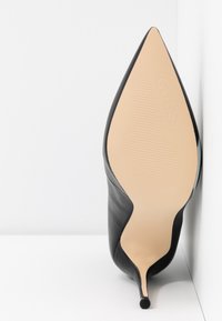 Chaussure à talon stiletto noir à bout pointu avec semelle beige embossée du mot "GUESS", positionnée verticalement contre un coin blanc.