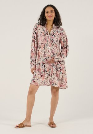 Femme enceinte debout, vêtue d'une robe fleurie longue jusqu'aux genoux à manches longues et de sandales marron, une main posée sur son ventre.