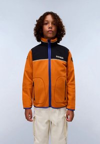 Noor juustega noor inimene, kellel on seljas oranž ja must lukuga fliisjakker ning beežid kotipüksid, seistes ühetoalise tausta ees.