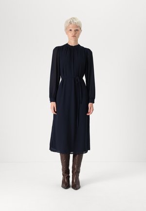 Robe longue bleu marine à manches longues avec un col rond, une taille à nouer et des détails froncés. Associée à des bottes brunes jusqu'au genou avec un design texturé.