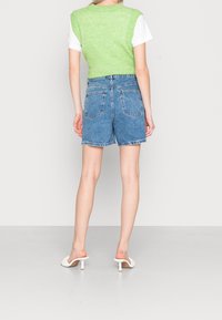Persona che indossa un maglione senza maniche verde chiaro sopra una camicia bianca, pantaloni corti in denim blu e mule bianche con tacco, in piedi contro uno sfondo semplice.