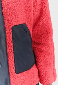 Czerwona polarowa kurtka z teksturowanym wykończeniem, z czarną kieszenią z tkaniny po lewej stronie i gładkim przednim panelem.