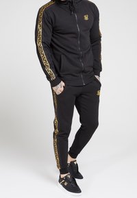 Felpa con cappuccio nera con zip e pantaloni jogger abbinati, caratterizzati da un motivo a catena dorata lungo i lati e dettagli con logo. Tessuto liscio e design aderente.