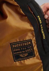 Zwarte jasinterieur met een gouden satijnen voering. Heeft een zwart label met gouden tekst "SUCCESSOR" en "PME LEGEND" merkbranding.