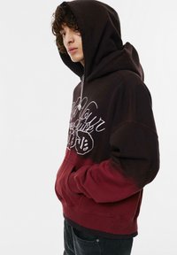 Mikina s kapucňou s tmavohnedým prechodom do burgundskej farby, vyšívaným textovým dizajnom a prednou klokanken. Mäkký materiál z bavlnenej zmesi.