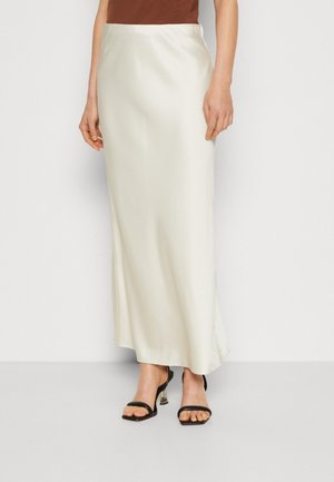 Femme portant une jupe maxi en satin couleur crème associée à des sandales noires à talons ouvertes et un haut marron.