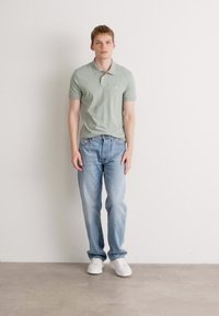 Lys grønn polo skjorte med korte ermer, kombinert med lyseblå jeans og hvite sneakers, stående mot en nøytral bakgrunn.