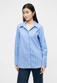 Γυναίκα που φοράει πουκάμισο με κάθετες ρίγες σε μπλε και άσπρο χρώμα, με φαρδιές μανσέτες, συνδυασμένο με σκούρο μπλε τζιν, στέκεται μπροστά σε λευκό φόντο.