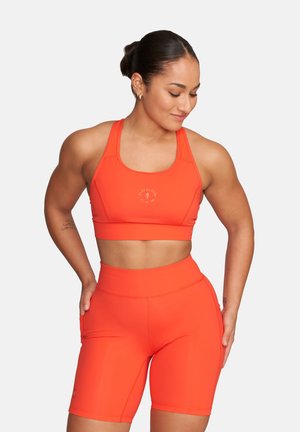 Kobieta w jaskrawo pomarańczowym sportowym staniku i wysokich krótkich spodenkach rowerowych, stojąca z rękami na biodrach i patrząca w dół, w lewą stronę.