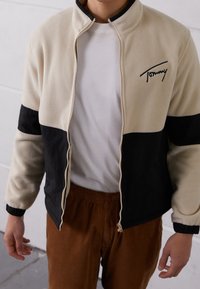 Fleecetakki kermaan ja mustaan, vetoketjulla suljettava, korkea kaulus ja kirjailtu logo. Takissa on kontrastiväriset hihat ja vyötärö.