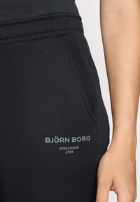 Svarta svettbyxor med en sidoficka och en tryckt logotyp i ljusgrön, med en slät textur och ett elastiskt midjeband.