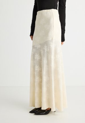 Naine, kellel on seljas pikk kreemikas lillelise pitsiga seelik, musta pika varrukaga särk ja mustad teravate ninadega kingad, seisab valgel põrandal.