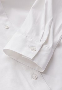 Nahaufnahme eines weißen Hemdes, das einen geknöpften Manschette mit weißen Knöpfen und glattem, strukturiertem Baumwollstoff zeigt. Sauberer Stich ist sichtbar.