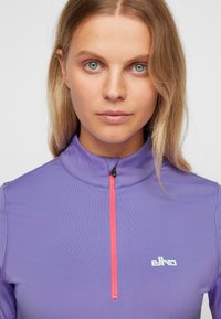 Lila langärmliges Sportoberteil mit hohem Kragen, halb im leuchtenden Pink durchgehend, und Logodetail in hellblau auf der Brust. Glatte Textur.