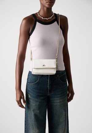 Borsa a tracolla bianca con un emblema triangolare, realizzata in materiale liscio. Presenta una tracolla a catena e una chiusura a patta, indossata su una maglietta rosa e jeans in denim.