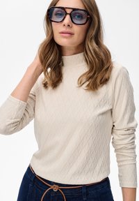 Vrouw met lichtbruin golvend haar, die oversized schildpadbril draagt, beige textuur lange mouw top en donkerblauwe spijkerbroek met een leren riem.