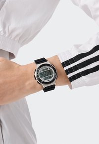 Digitális óra fekete gumi szíjjal és ezüst színű fém házzal, nagy digitális kijelzővel, amelyen az idő, dátum és stopper funkciók találhatók.