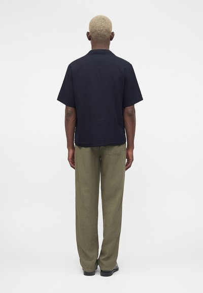 Άτομο με κοντά ξανθά μαλλιά στέκεται με την πλάτη, φοράει μαύρο κοντομάνικο πουκάμισο, χακί παντελόνι και μαύρα παπούτσια σε απλό φόντο.