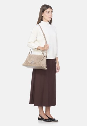 Borsa in suede beige con patta curva, hardware in argento e due opzioni di trasporto. Texture liscia, abbinata a un maglione leggero e una gonna marrone.