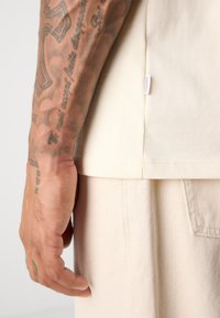 Un braccio tatuato poggiato accanto a una camicia color crema e pantaloni coordinati con cuciture visibili e un piccolo etichetta in tessuto etichettata "LINDBERGH".
