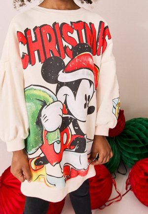 Enfant portant un sweat blanc surdimensionné avec Mickey Mouse en bonnet de Père Noël tenant un sac vert, avec « NOËL » en lettres rouges majuscules au-dessus.