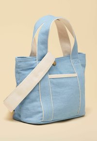 Sac fourre-tout en toile bleu clair avec des anses crème et une poche avant, sur fond beige.