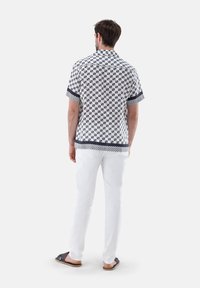 Camicia a maniche corte bianca con un motivo geometrico blu, con colletto e rifiniture a contrasto. Abbinata a pantaloni bianchi e sandali casual.