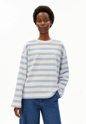 Femme aux cheveux courts et bouclés portant une chemise blanche et bleu clair rayée à manches longues et un jean bleu, debout avec une main dans la poche.