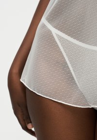 Povečani posnetek boku in roke osebe, ki nosi prozorno belo mrežasto spodnje perilo z vzorcem pik in trdim robom.