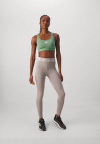 Zielony stanik sportowy z białym logo, jasnoszare legginsy z białymi detalami w pasie i czarne buty sportowe. Gładka tekstura materiału.