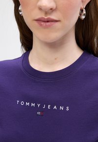 Violets kokvilnas T-krekls ar apaļu kakla izgriezumu, kuram ir "TOMMY JEANS" logotips baltā krāsā un nelielas krāsainas akcenti zem tā. Gluda tekstūra.