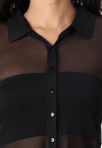 Lähikuva henkilöstä, joka käyttää mustaa läpinäkyvää nappipaitaa, jonka alla on kiinteä musta bandeau.