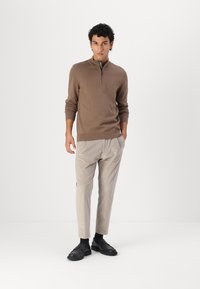Pull en maille marron zippé avec manches longues, associé à un pantalon beige clair ajusté et des chaussures noires, sur fond blanc uni.
