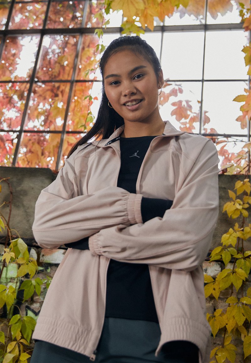 Veste de sport rose clair avec des poignets élastiques, portée sur une chemise noire arborant un logo, sur fond de feuillages d'automne vibrants.