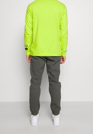 Camisa de manga larga de color verde neón con detalles en negro en las mangas, combinada con pantalones jogging grises y zapatillas blancas, mostrada desde la parte posterior.