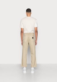 Muž stojící zády, oblečený v béžových kalhotách, bílých teniskách a béžovém tričku s texturovaným bílým rukávem na pravém rameni.