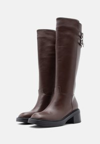 Καφέ δερμάτινες μπότες μέχρι το γόνατο με στρογγυλεμένη μύτη, χοντρό τακούνι και δύο ασημένιες αγκράφες στην εξωτερική πλευρά. Λείο υλικό με ραφές για διακόσμηση.