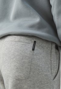 Grå hettegenser kombinert med grå joggebukser. Buksene har en sidelomme og en subtil svart merkelapp på livet. Myk stofftekstur.