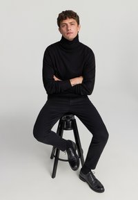 Czarny sweter z wysokim kołnierzem o prążkowanej fakturze, w połączeniu z czarnymi spodniami slim fit i czarnymi sznurowanymi butami, siedzący na czarnym stołku.