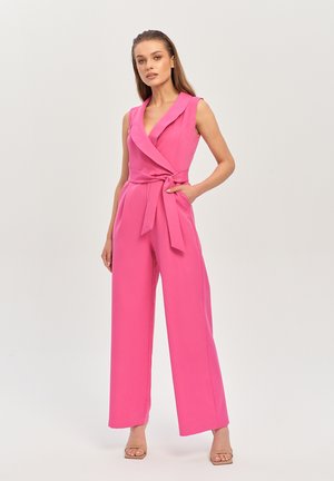 Kobieta w bezrękawnym, jaskrawo różowym kombinezonie z szerokimi nogawkami i paskiem w talii, stojąca z jedną ręką w kieszeni na jednolitym tle.