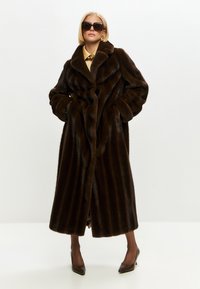 Brun faux fur frakke med sjalskrave, lodret ribbet tekstur og fuldlængde silhuet, parret med mørke spidse hæle.