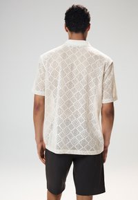 Chemise blanche à manches courtes en crochet avec des découpes en forme de losange et un col, associée à un short foncé, vue de dos.