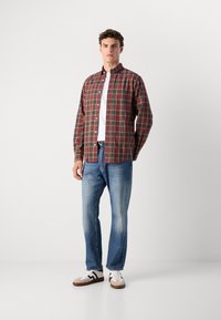 Geruite overhemden in rood, groen en gele tinten over een wit shirt, gecombineerd met blauwe jeans en witte sneakers met bruine accenten.