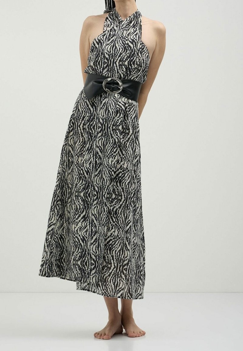 Femme debout pieds nus dans une robe longue sans manches à motifs noir et blanc avec une large ceinture noire avec une grande boucle ovale.